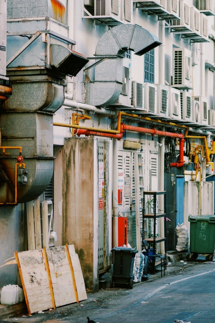 Back alley view with air conditioning units, industrial pipes, and urban clutter, showcasing city life.